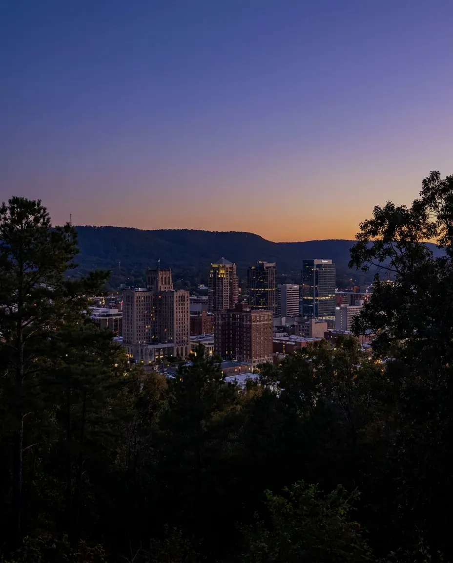 A downtown of stone and brick towers below a wooded ridge at dusk.