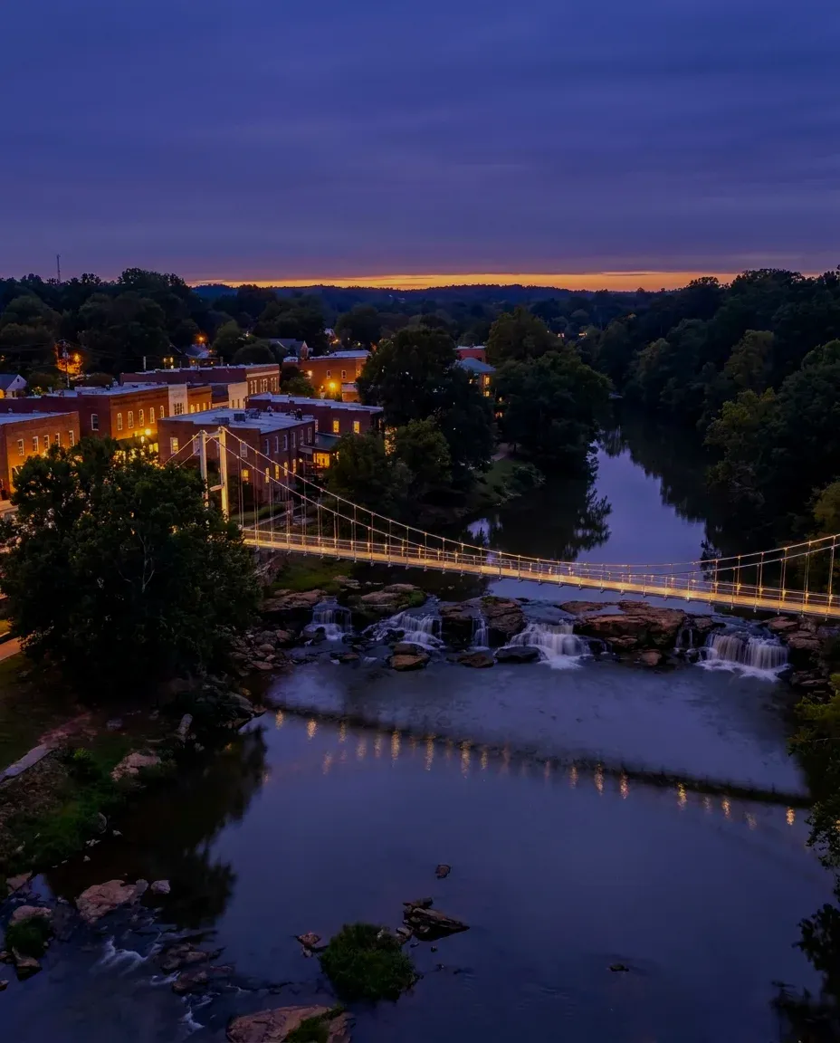 A footbridge over low waterfalls in a leafy downtown at dusk.