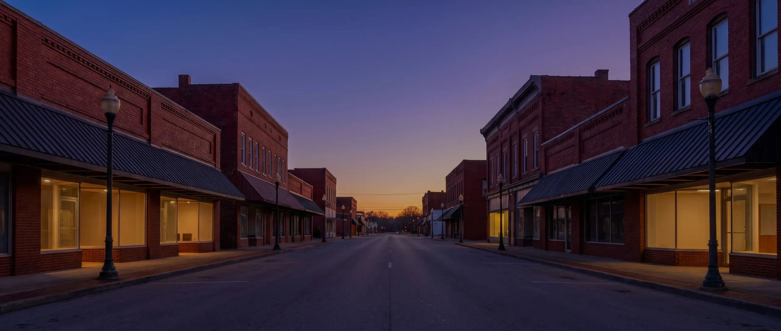 A small-town main street of brick buildings with lit shop windows at dusk.