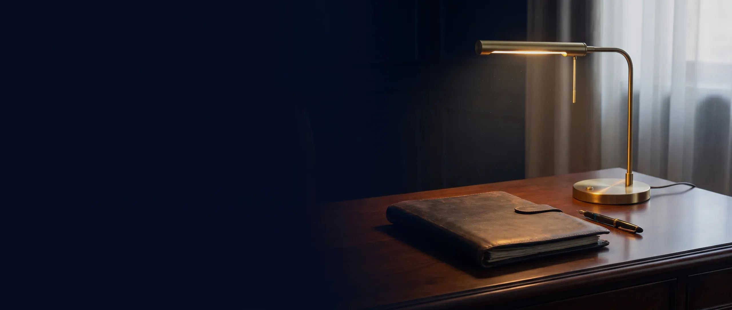 A closed leather folio, a fountain pen and a brass desk lamp on a dark desk.