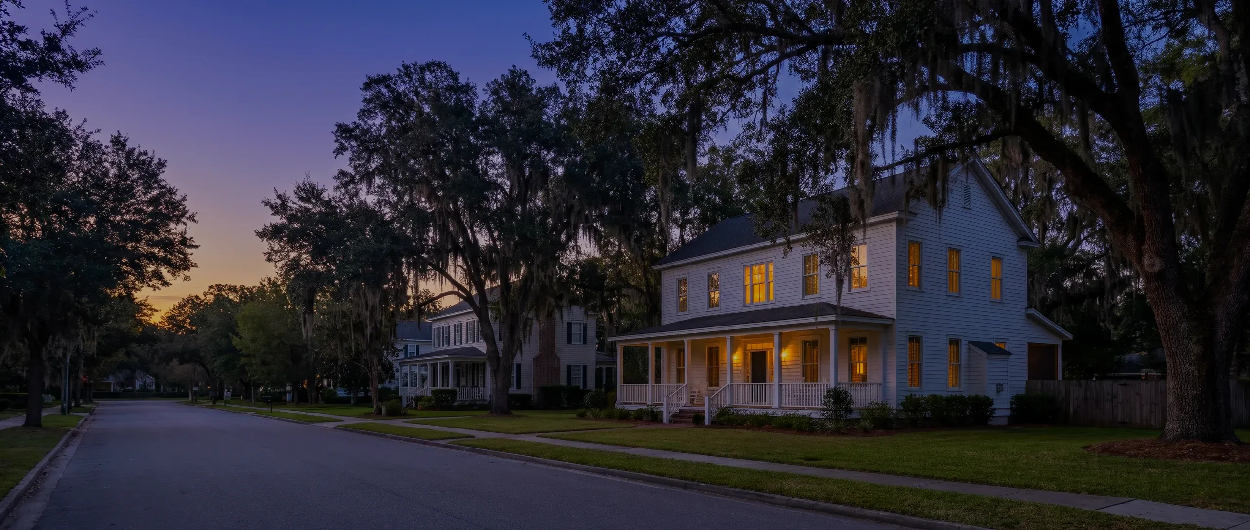 A tree-lined street at dusk, a white house with a lit front porch under live oaks.